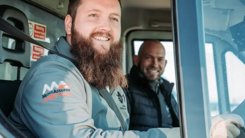 Bearded Bros crew member wearing a Move Assured branded jacket, smiling from inside a removals van cab with a colleague